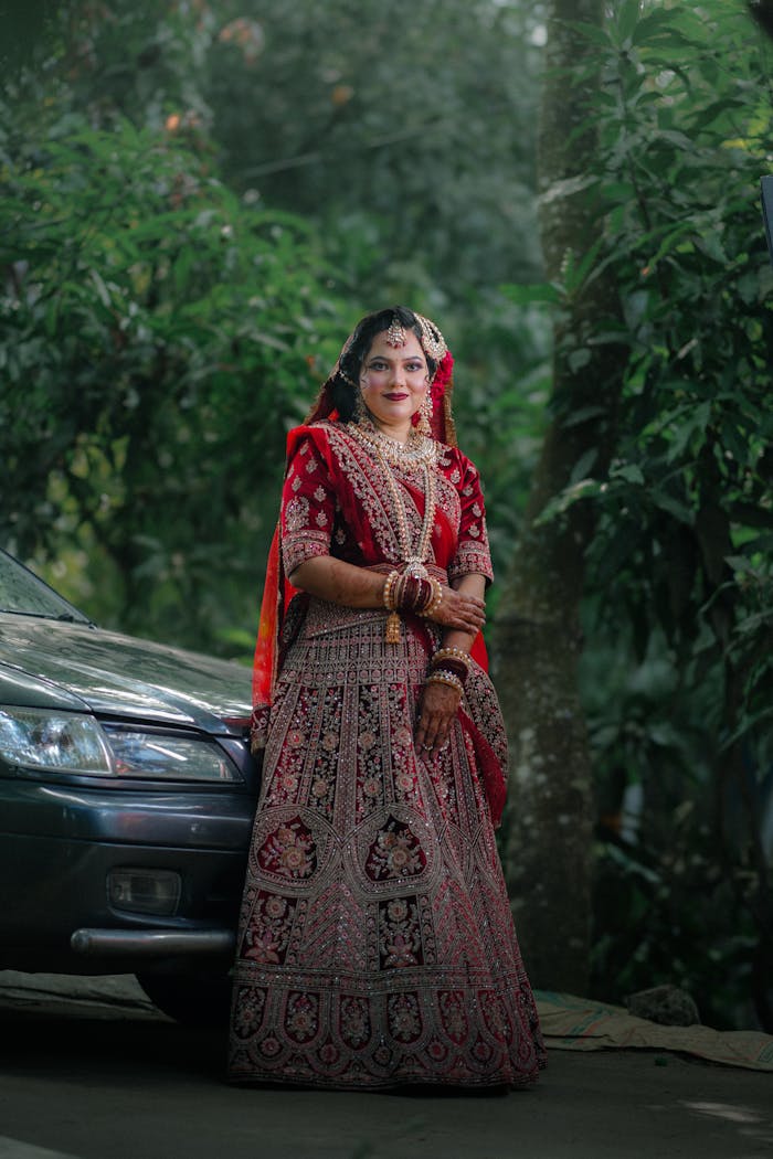 A beautiful bride in traditional Indian attire stands gracefully outdoors beside a car.